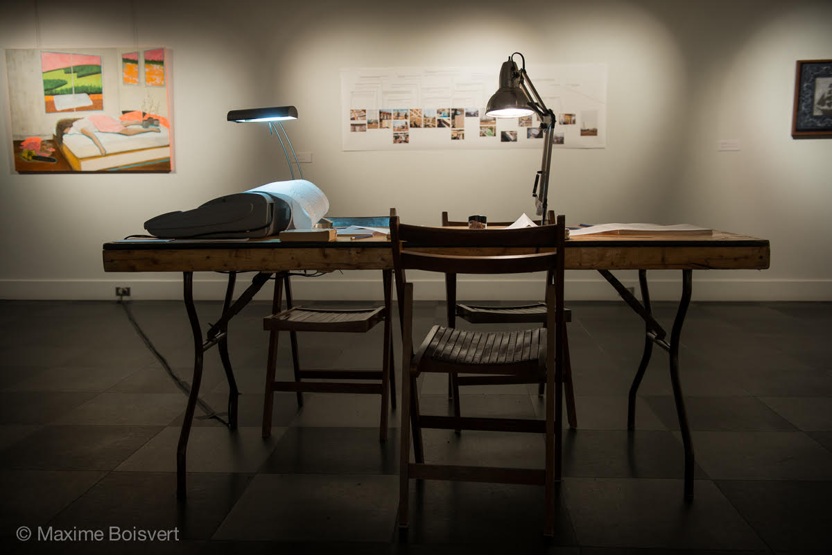 An art gallery with works on the walls and a wooden folding table with three wooden folding chairs around it. On the table is a typewriter printing poetry on continuous-feed paper underneath a desk lamp, surrounded by books of poetry and writing utensils for people in the gallery to annotate its writings